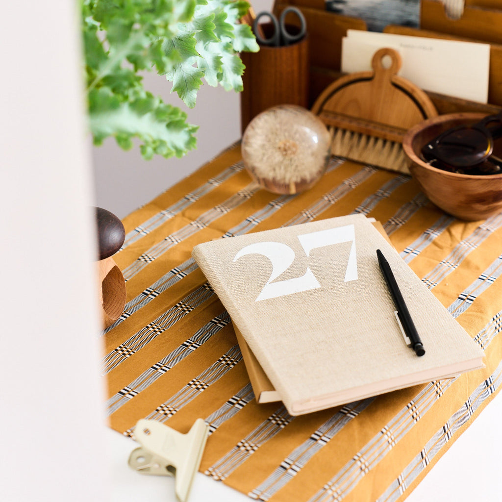 A desk scene with a natural linen 2027 Diary planner, a black pen, a striped yellow-and-white runner, a wooden organizer, a plant, and other desk accessories.