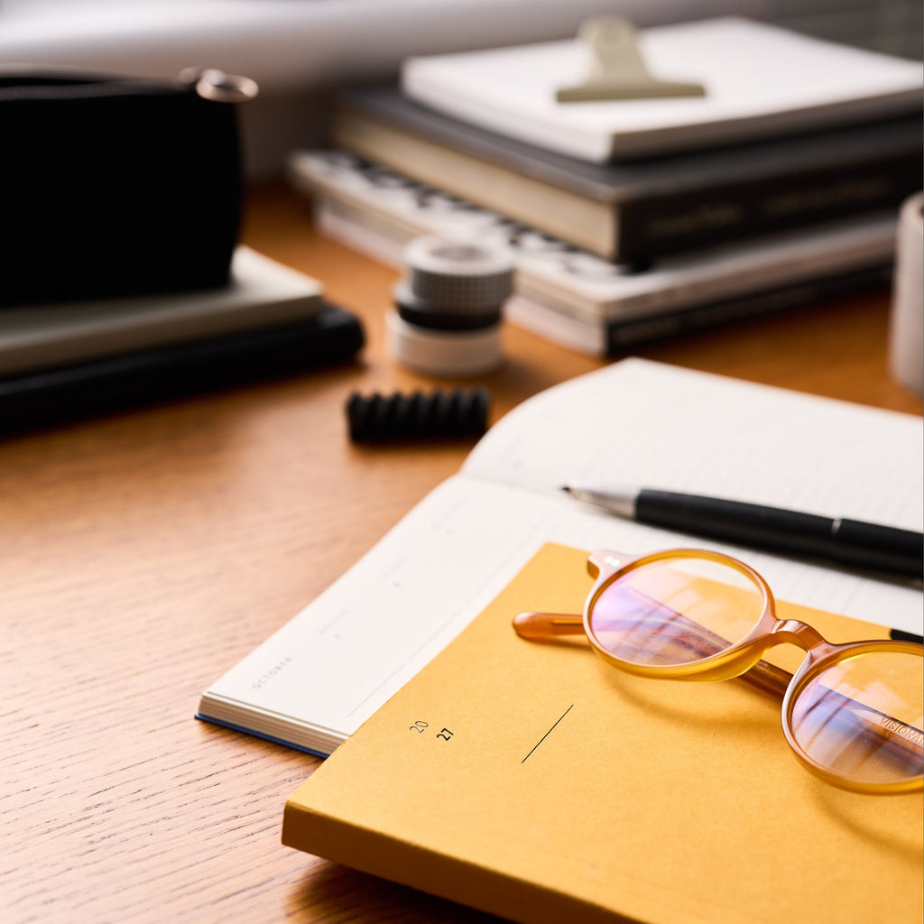 Round amber eyeglasses resting on a mustard yellow 2027 diary on a wooden desk, surrounded by books, a pen, and office items in a softly blurred workspace setting.
