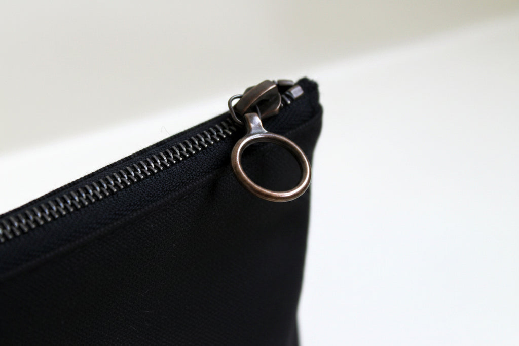 Close-up of a black canvas pencil case with a brass zipper and metal ring on a light background